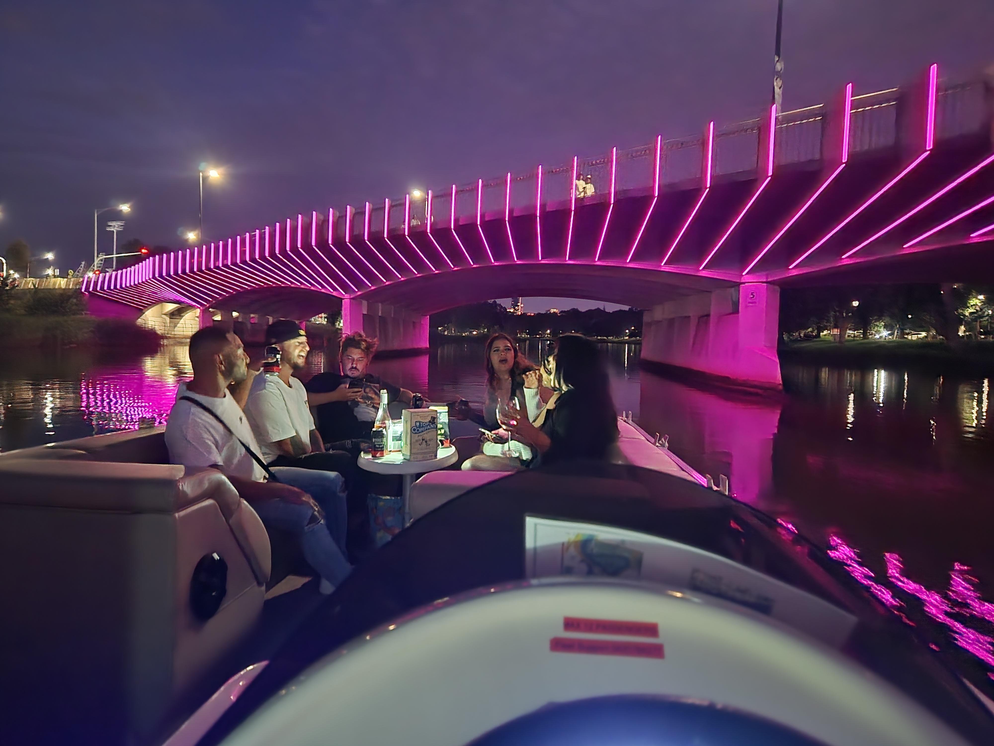 A pontoon boat passing under a bridge lit pink at night