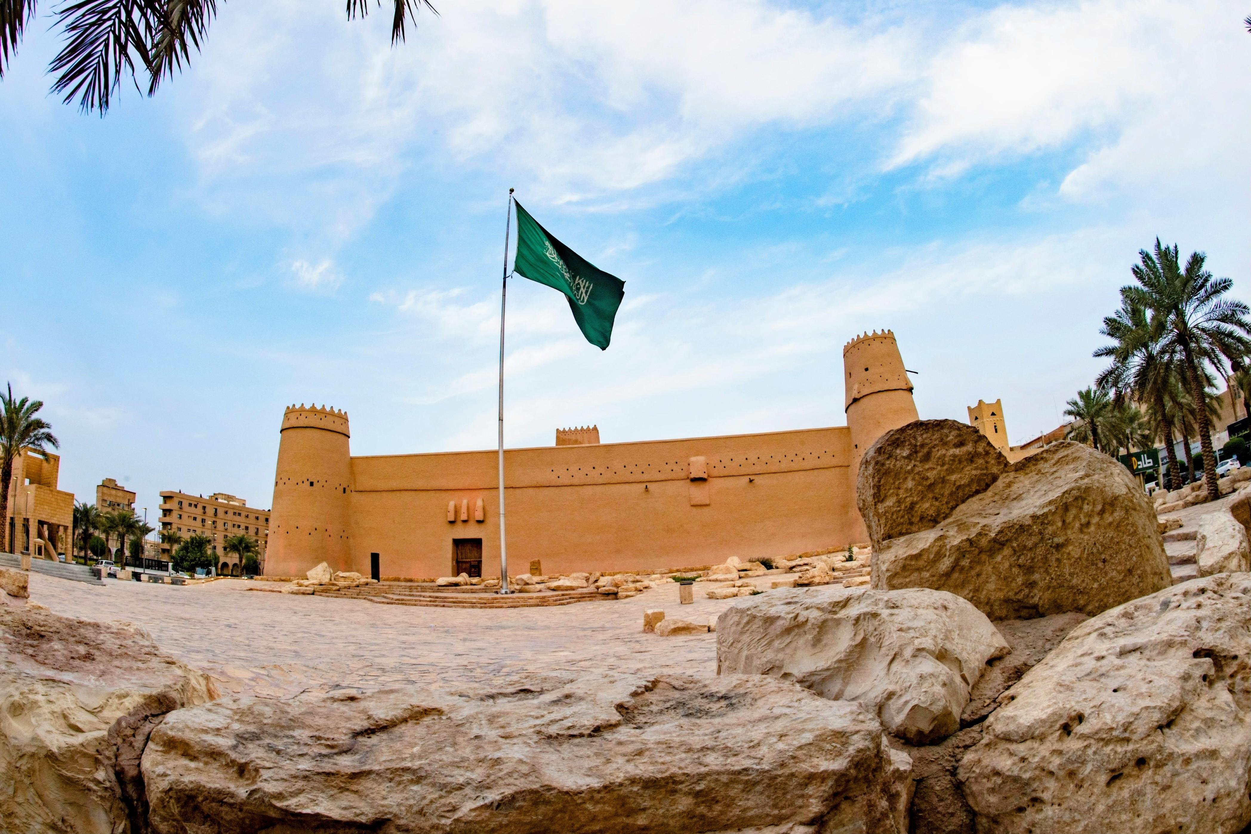 The mud-brick walls and round tower of the Masmak Fortress in old Riyadh, with palm trees and the Saudi flag