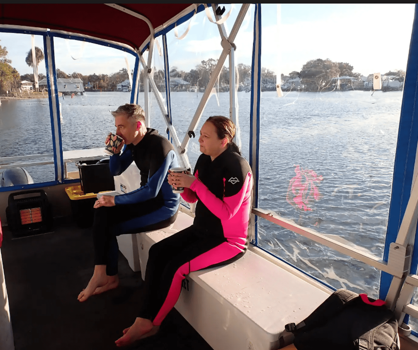 Two guests in wetsuits warming up with drinks aboard a covered boat