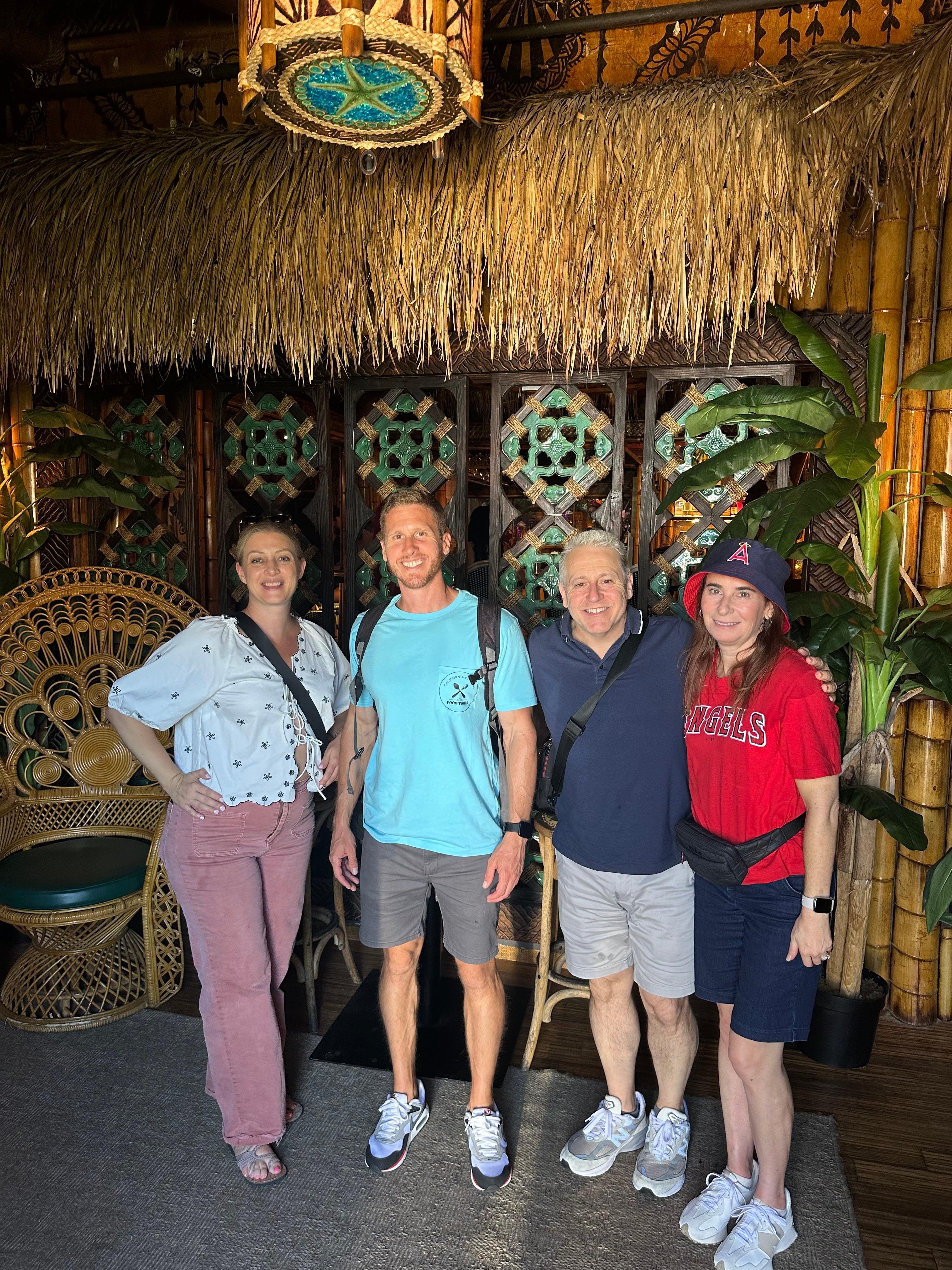 Four people, two women and two men, smile while posing arm-in-arm in front of a tiki-themed bar entrance with a thatched grass roof, bamboo pillars, green decorative tiles, rattan peacock chairs, and tropical plants.