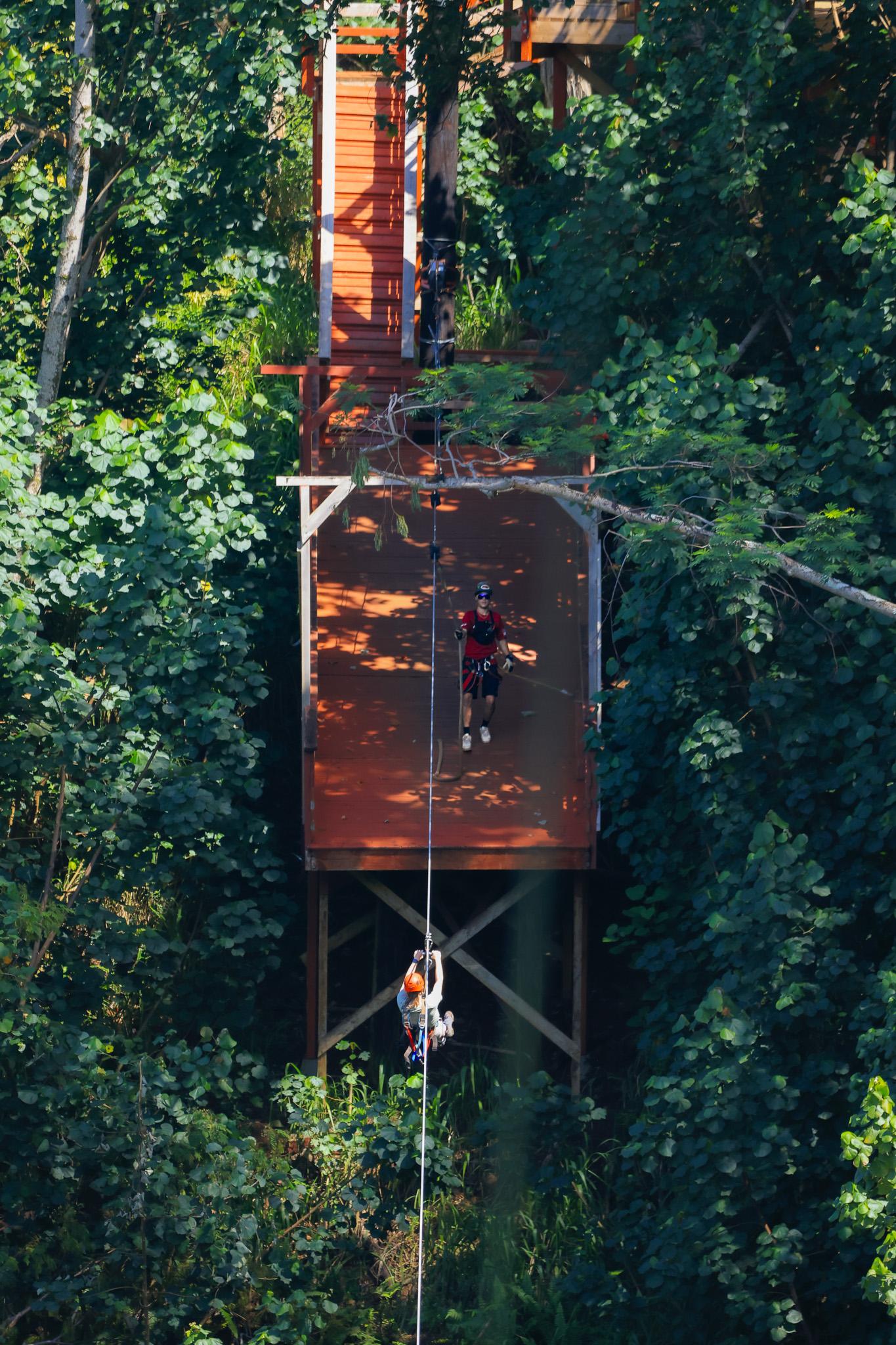 Two riders on a zipline beside a tall wooden course structure