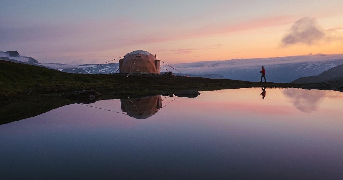 Et kuppeltelt på en bjergtop ved et stille tjern ved solnedgang, med en person på stien ved siden af