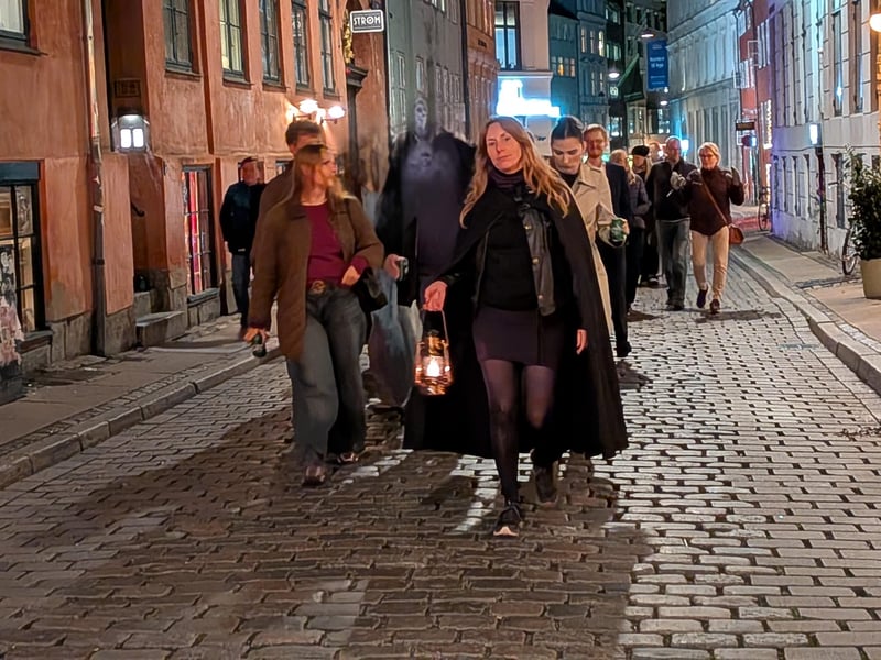 Kvinde i sort frakke går med en lanterne i hånden ned ad en brostensbelagt gade i København, med forbipasserende bag sig