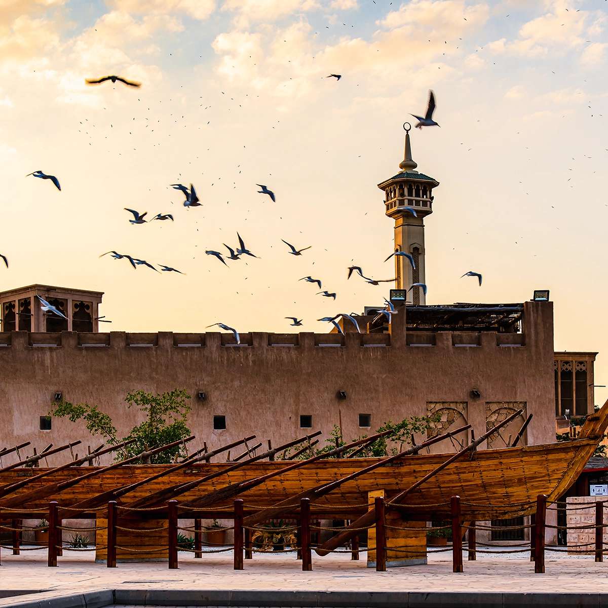 Traditional wooden dhow boats in front of Al Fahidi Fort, with a minaret and flock of birds at sunset.