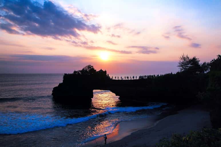 A scenic view of a sunset over the ocean with a rocky archway and trees.