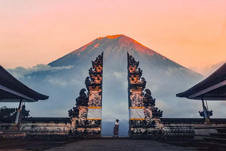 A person stands between ornate gates with a mountain in the background at sunset.