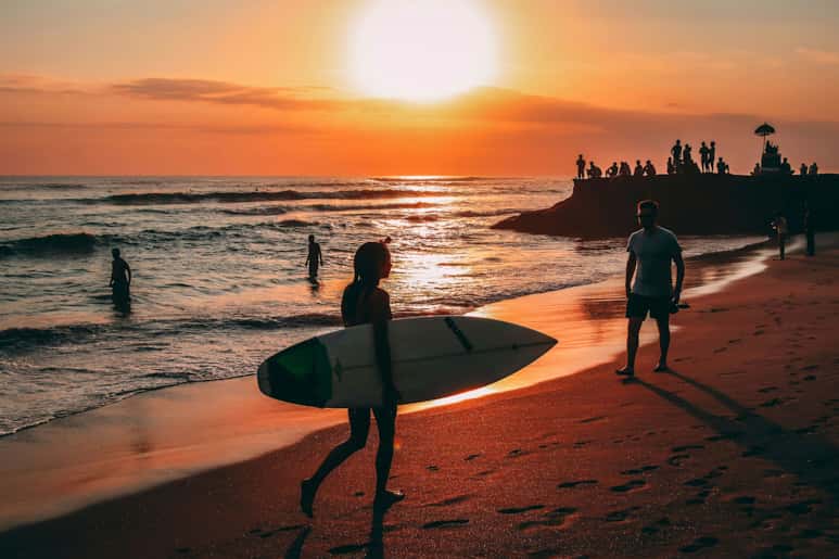 A surfer walking along the beach at sunset with people in the background.