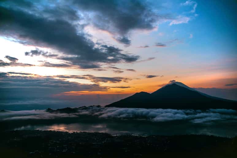 A scenic view of a mountain at sunset with clouds and a lake below.