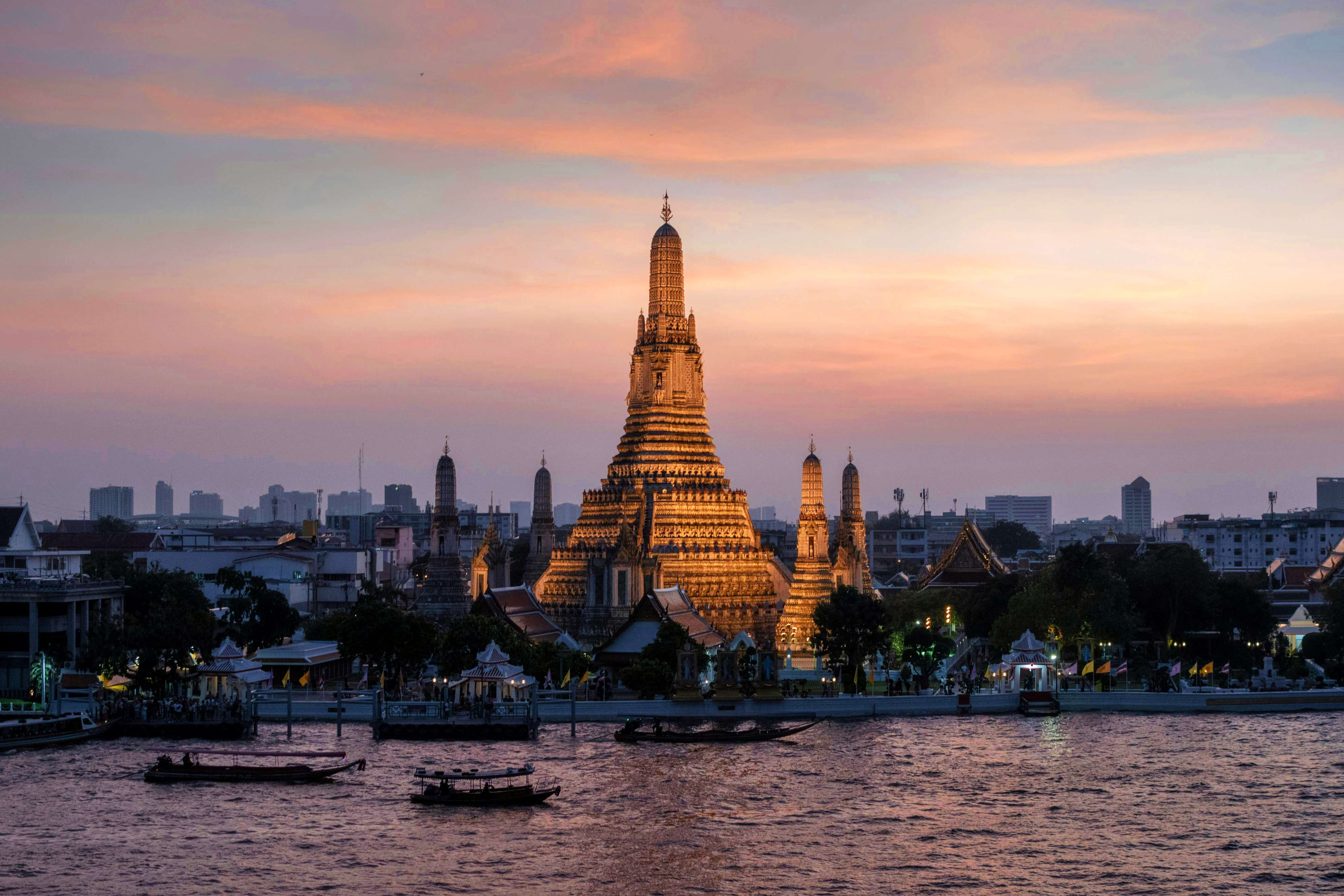 Candi Wat Arun bersinar keemasan saat matahari terbenam dengan awan merah muda dan biru di latar belakang