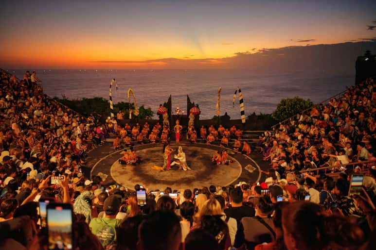A large crowd watches a traditional performance at sunset by the ocean.