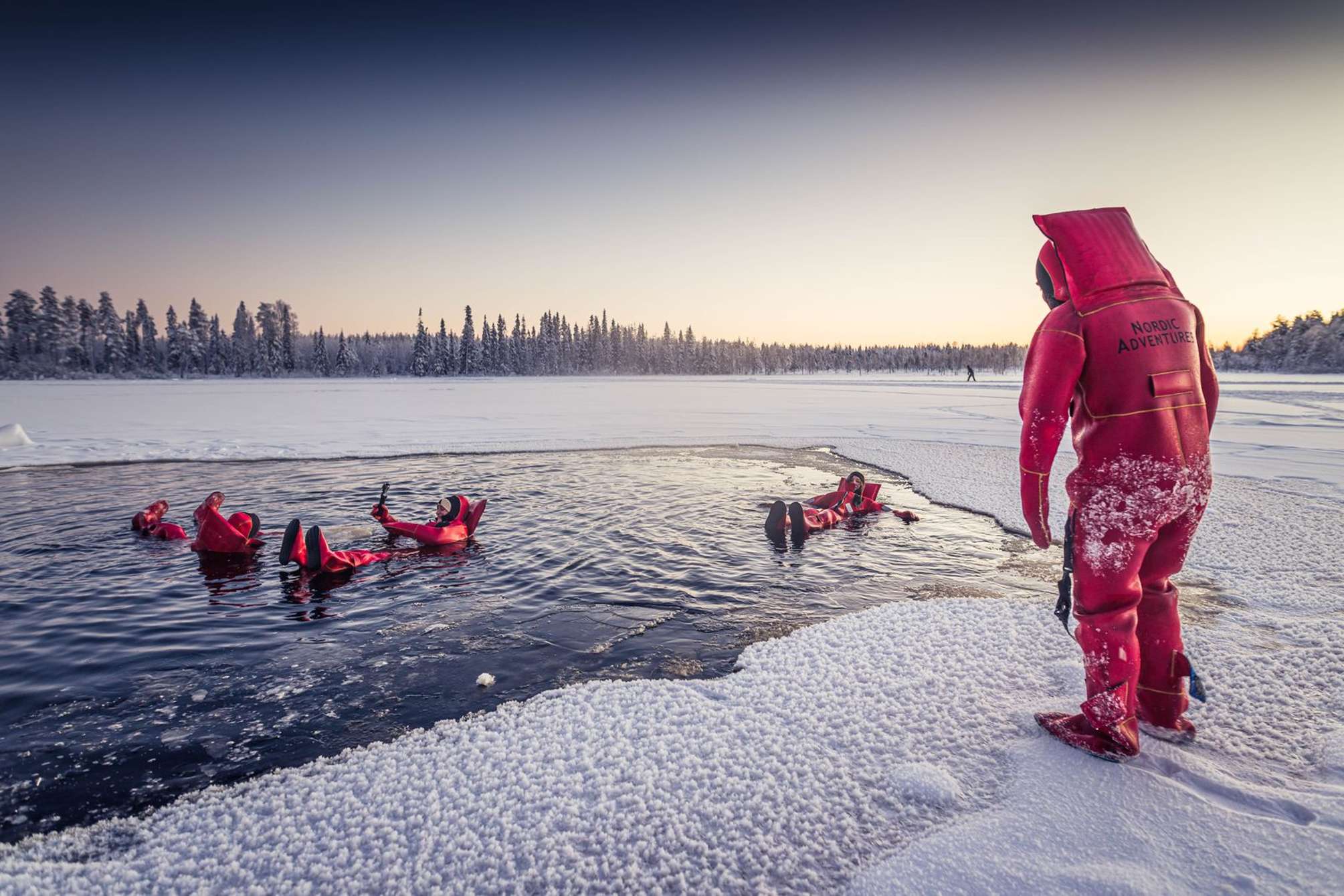 Floating auf einem zugefrorenen See bei Rovaniemi mit Nordic Adventures: Menschen im Überlebensanzug treiben auf dem Rücken im eiskalten Wasser Lapplands, rundherum Schnee, danach beheizte Hütte