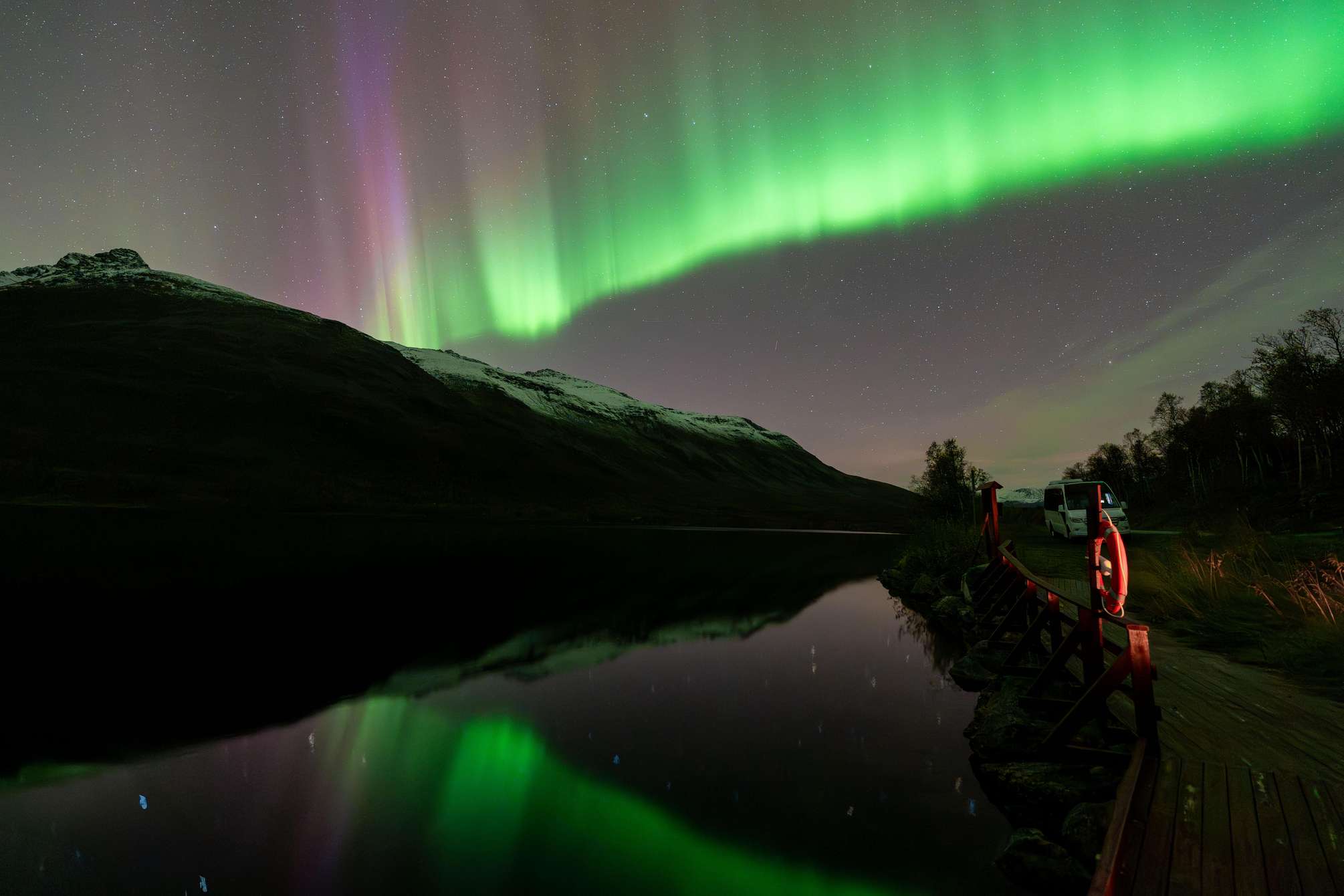 Nordlichtjagd mit Northern Norway Travel bei Tromsø: Lagerfeuer im Schnee unter grünem Himmel, Teilnehmende im Thermoanzug mit warmem Essen und Getränken
