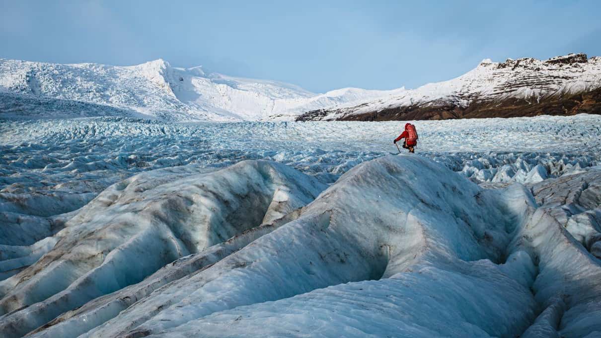 A lone hiker in a red jacket crossing the ridged blue ice of a Vatnajökull outlet glacier, with snow-covered peaks rising behind