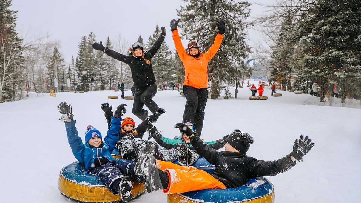 A family posing on colorful snow tubes with arms raised among snowy pines at Valcartier