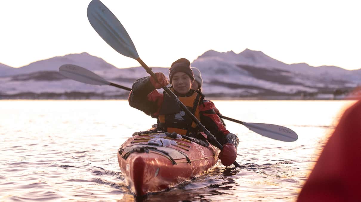 A kayaker in a red sea kayak paddling toward the camera with snowy mountains behind at sunset