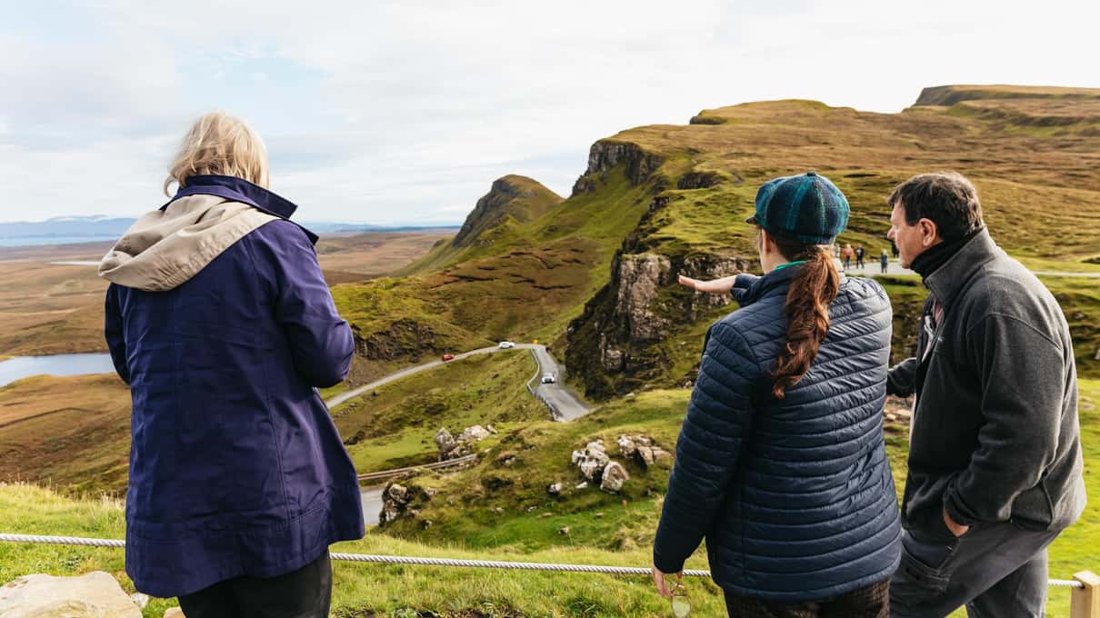 Small group at a Quiraing viewpoint on the Isle of Skye, a guide gesturing toward the road winding below the green landslip ridges