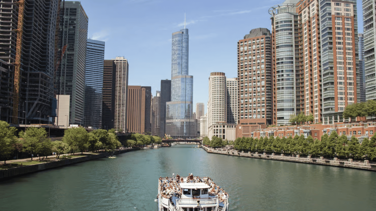 A Wendella sightseeing boat full of passengers on the green Chicago River between glass towers and tree-lined banks