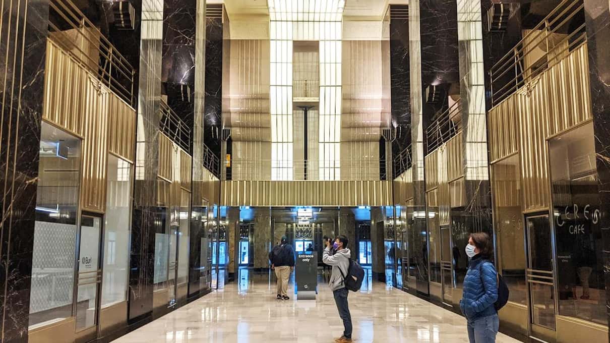 背の高いエレベーターの扉がある、磨かれた石のアールデコのロビーにいる少人数のツアーグループ