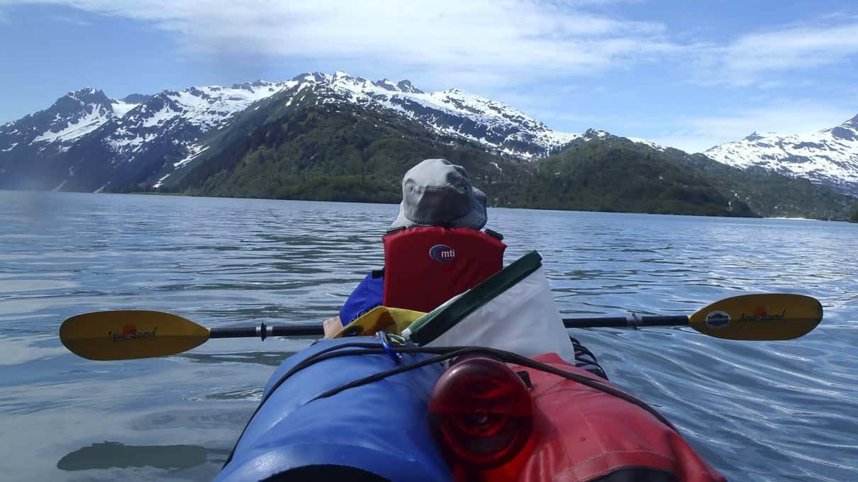 View over the bow of a tandem kayak toward snow-streaked mountains ringing a flat calm inlet