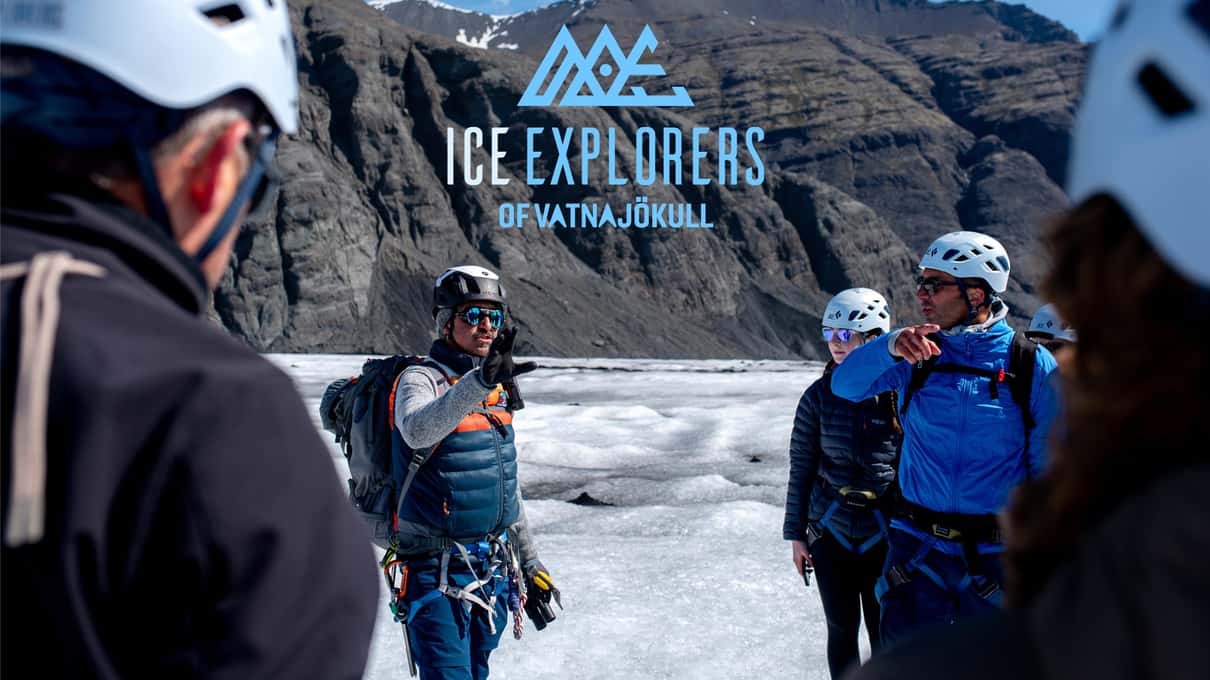 Helmeted guests gathered around a guide on a grey glacier surface, dark mountain walls rising behind them
