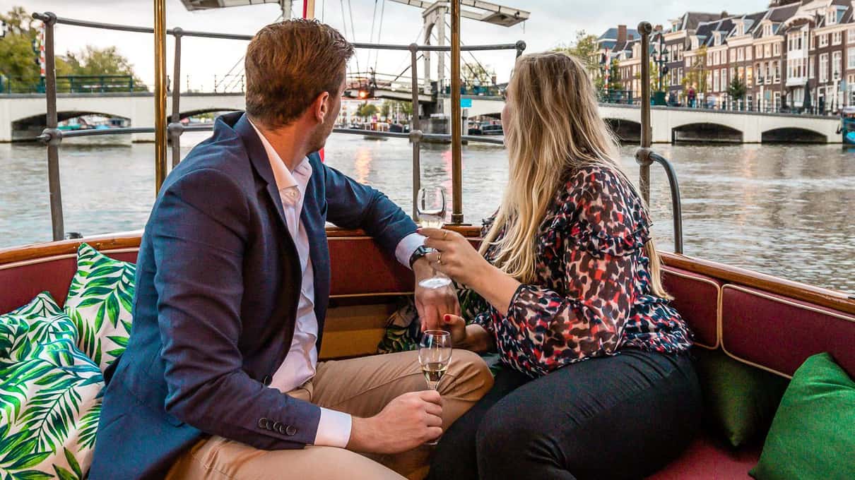 Pareja sentada en un barco con cojines, con el Magere Brug y casas del Amstel al fondo