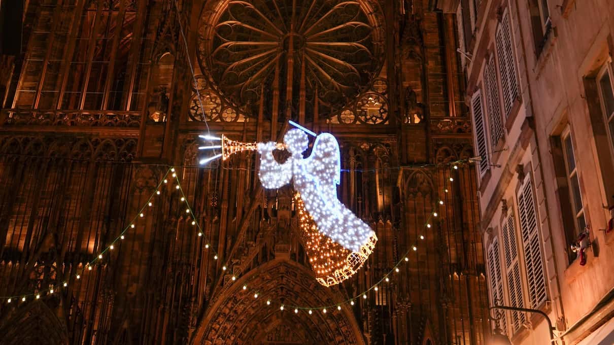 The carved sandstone facade of Strasbourg cathedral at night with a glowing angel of white lights hung across the lane in front