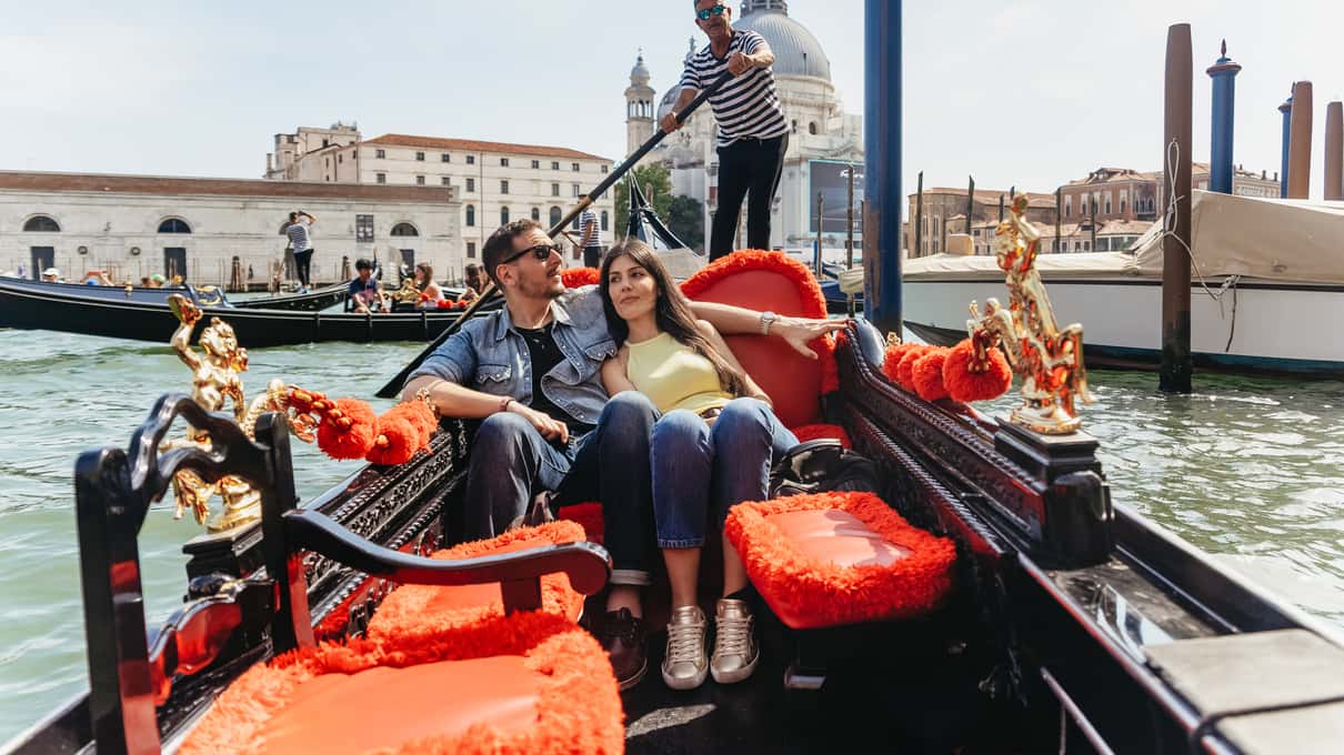 Paar sitzt auf roten Polstern in einer Gondel auf dem Canal Grande, hinter ihnen rudert der Gondoliere, im Hintergrund die Kuppel der Salute-Kirche