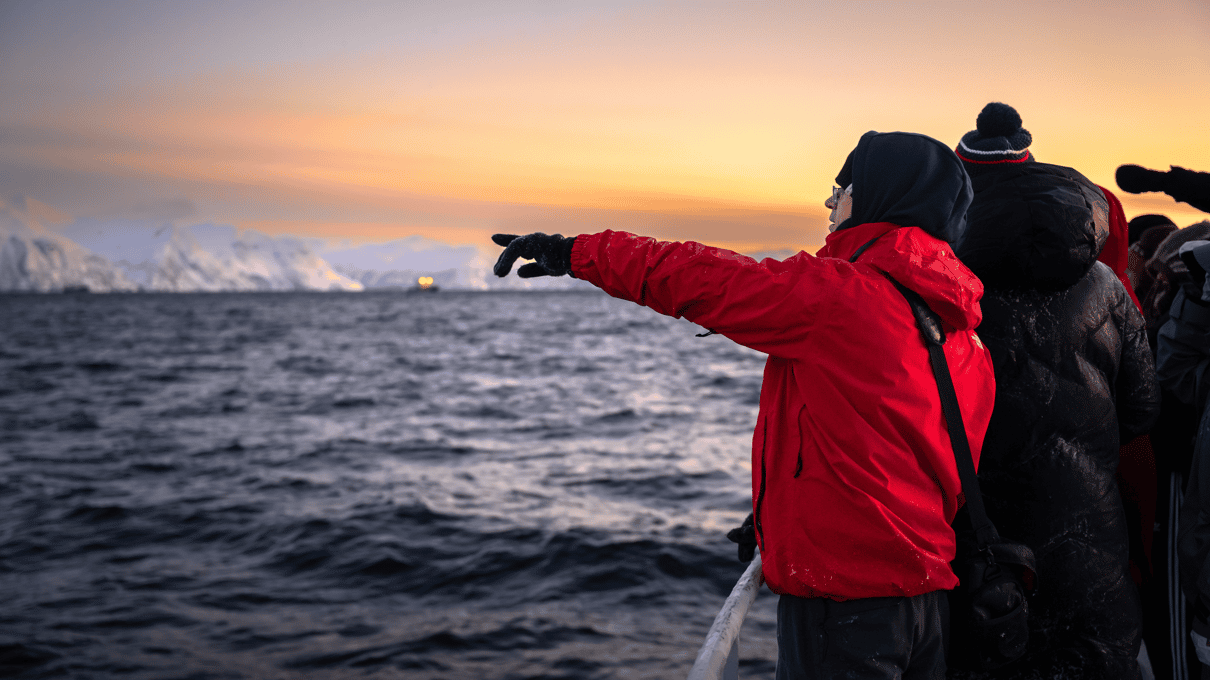 Guests in winter jackets on the deck of a boat, one in red pointing out to sea in the orange winter light