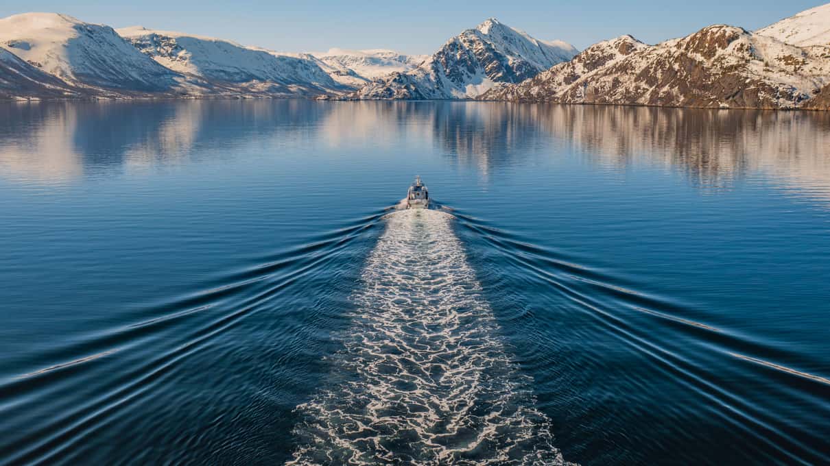 A small boat drawing a long white wake down a calm fjord between snow-covered mountains