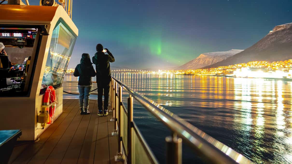 Two passengers on the deck of a boat looking at a faint green aurora above the lit-up Tromsø waterfront