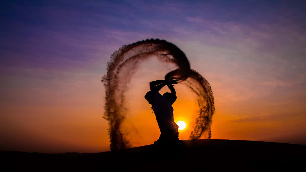 Silhouette di una persona che lancia la sabbia in un grande arco contro il sole che tramonta sulle dune