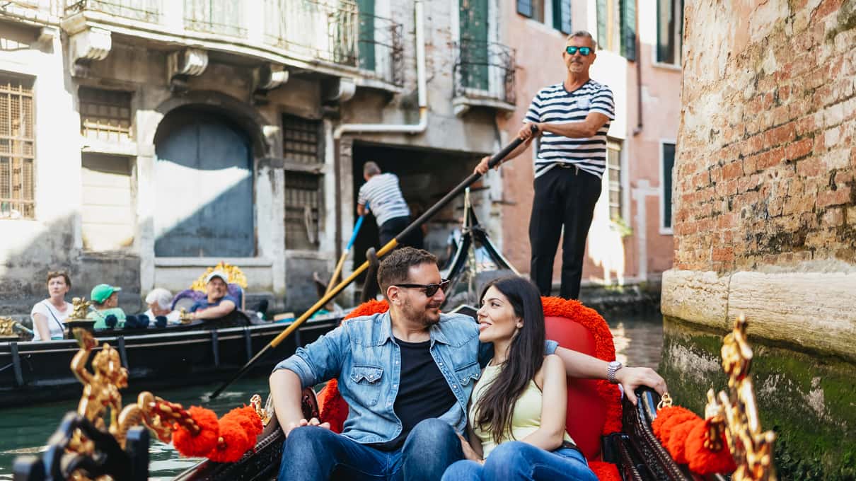 Couple souriant assis sur les coussins rouges d’une gondole dans un canal étroit, derrière eux le gondolier en marinière rame debout