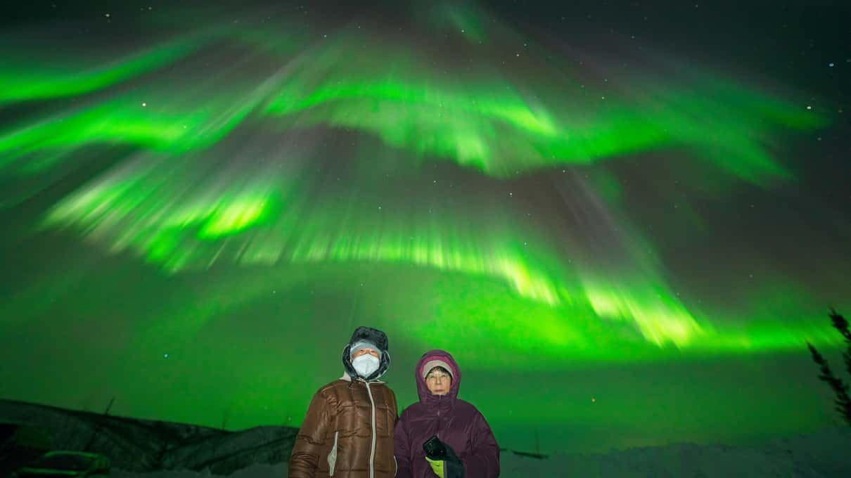 两位客人在雪地上摆姿势,头顶是横跨夜空的鲜绿色极光带
