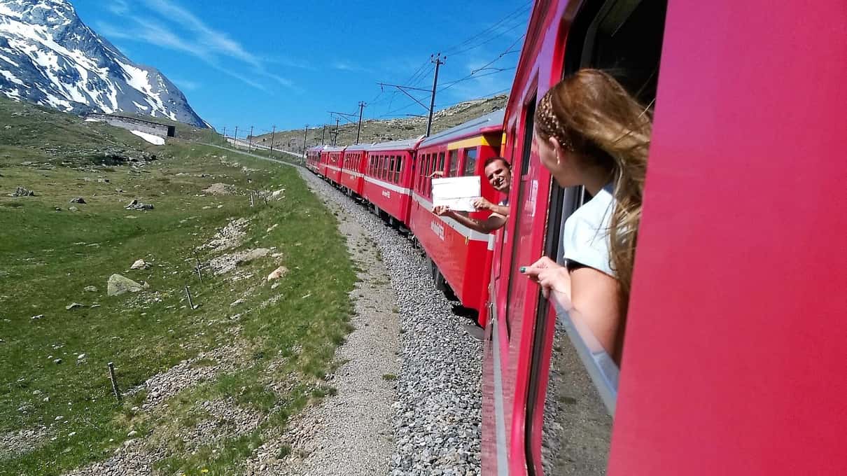 Travelers leaning out of the open windows of the red Bernina train on a grassy mountain stretch