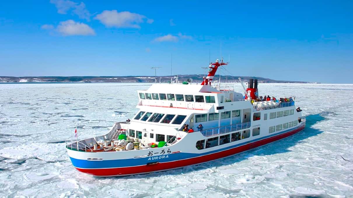 The Aurora icebreaker with its red hull and white decks moving through drift ice under a blue sky