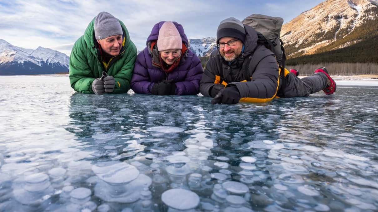 Three people in winter jackets and hats lying side by side on the ice, looking down at the bubbles