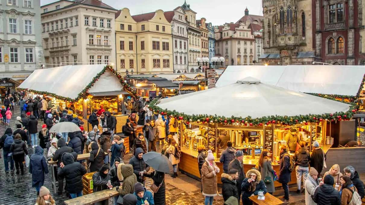 Wooden Christmas market huts on Old Town Square in daylight, with crowds of visitors walking between the stalls
