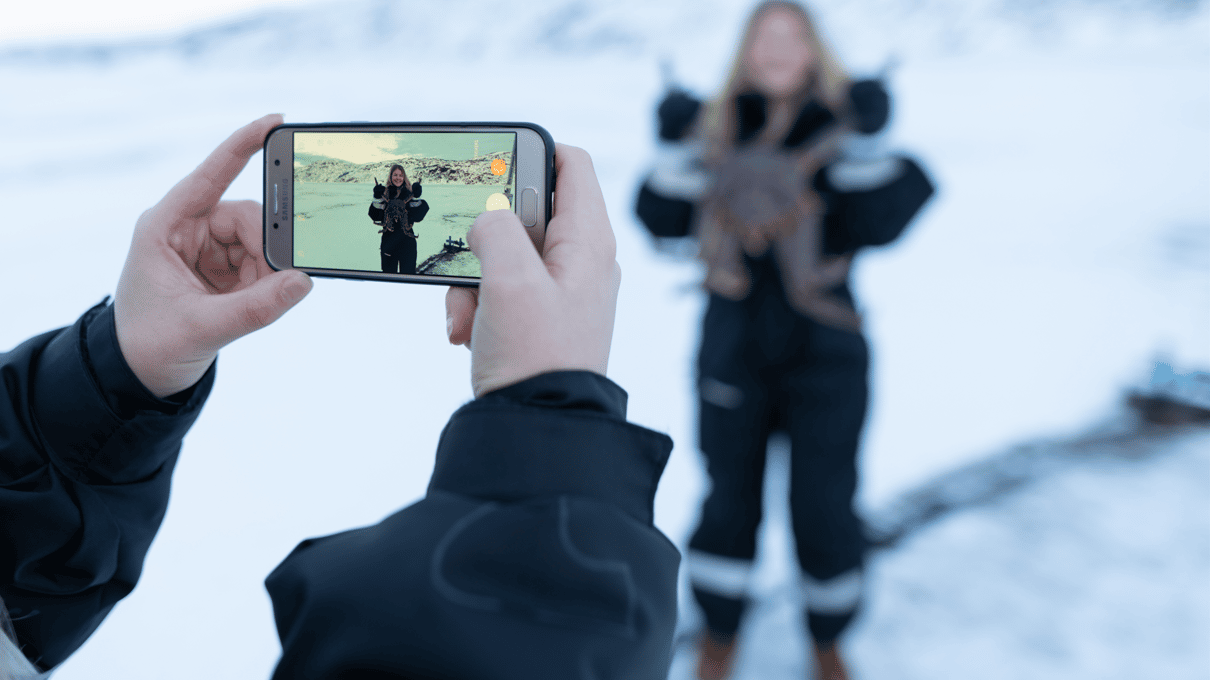 A phone taking a picture of a guest in a black thermal suit holding a red king crab out on the frozen fjord