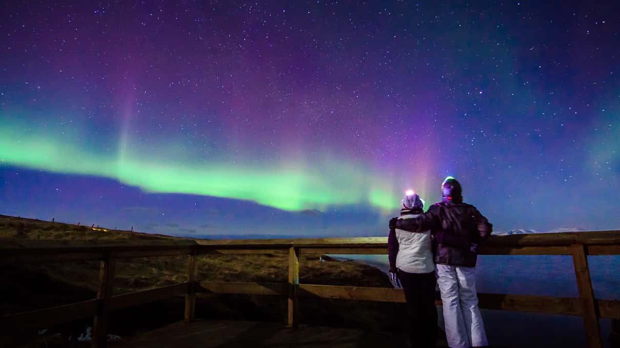 Paar an einem Holzgeländer unter violettem und grünem Polarlicht über einer isländischen Bucht