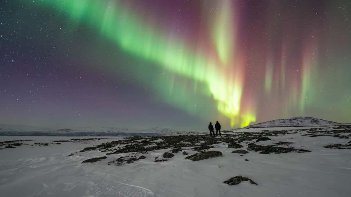 Two people standing on a snowy lake shore under green and purple aurora curtains