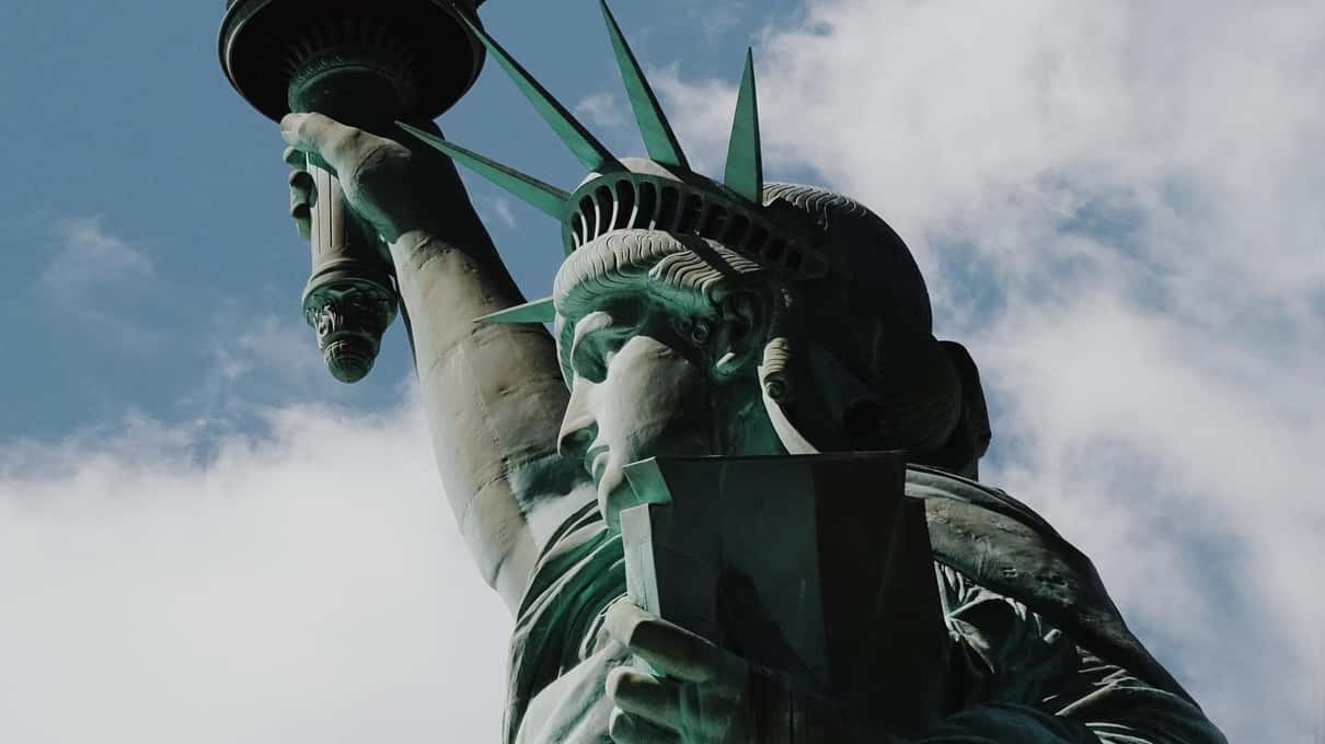 The Statue of Liberty's crown, face, tablet and raised torch arm seen from below against a cloudy sky