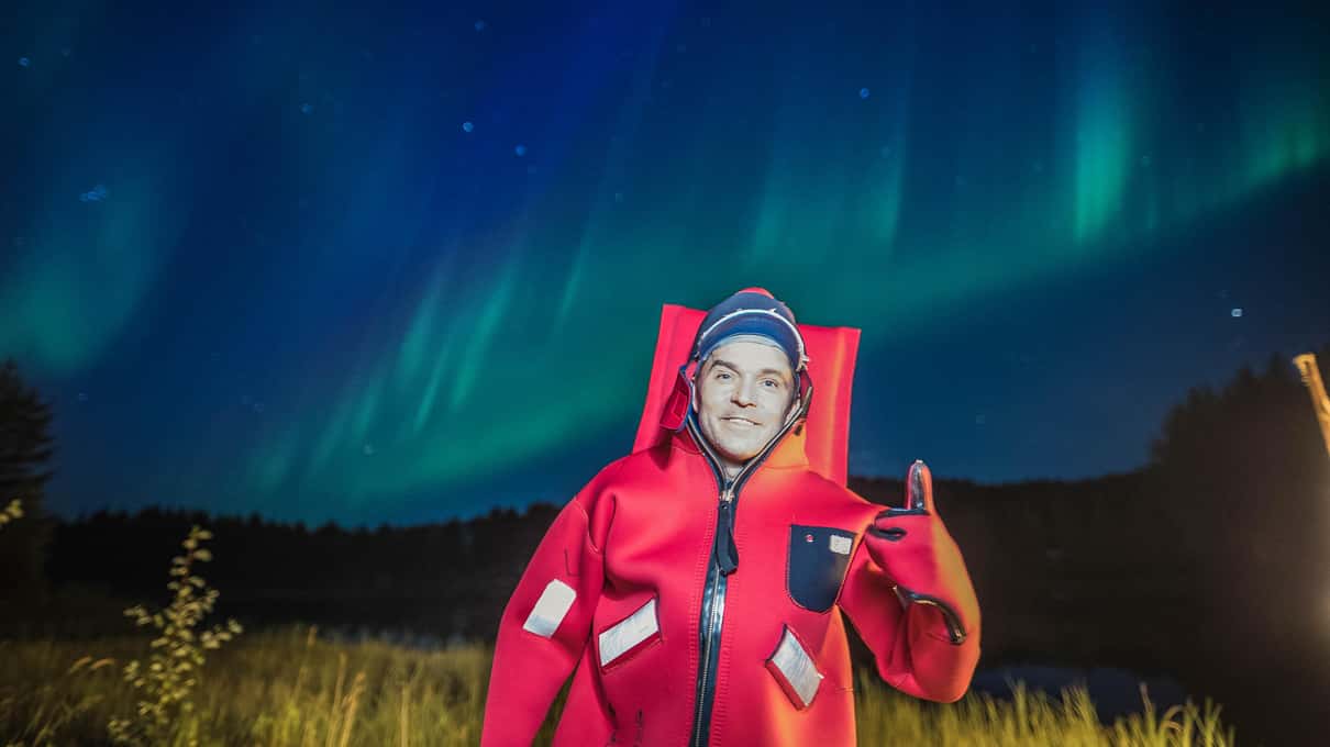 A floater in a red rescue suit giving a thumbs up in the ice hole under green northern lights