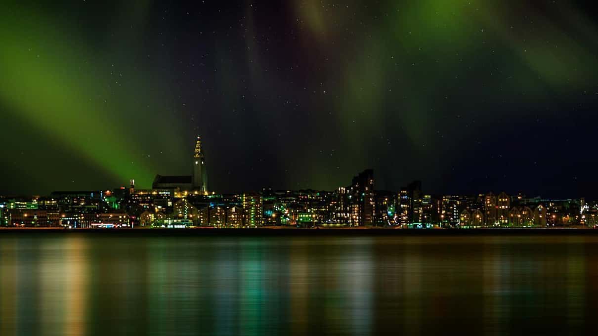 Reykjavik's skyline seen from Faxaflói Bay with a green aurora arching above the city lights