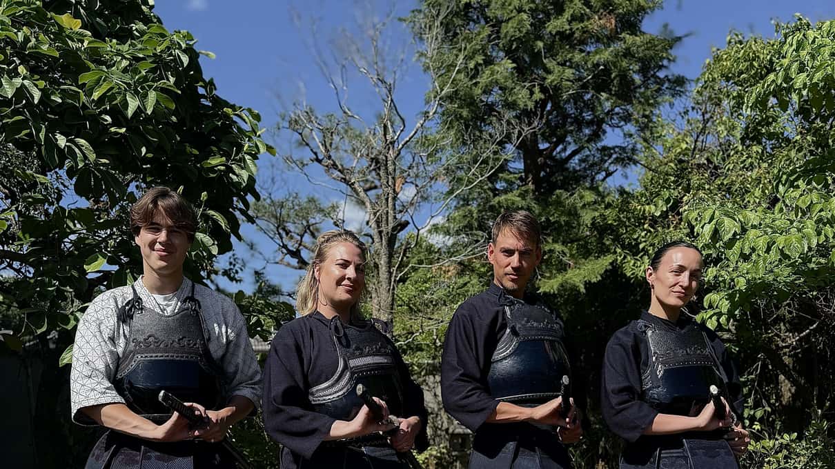 Čtyři hosté v modrých uniformách kenda drží své přilby na slunné zahradě v Kendo Experience