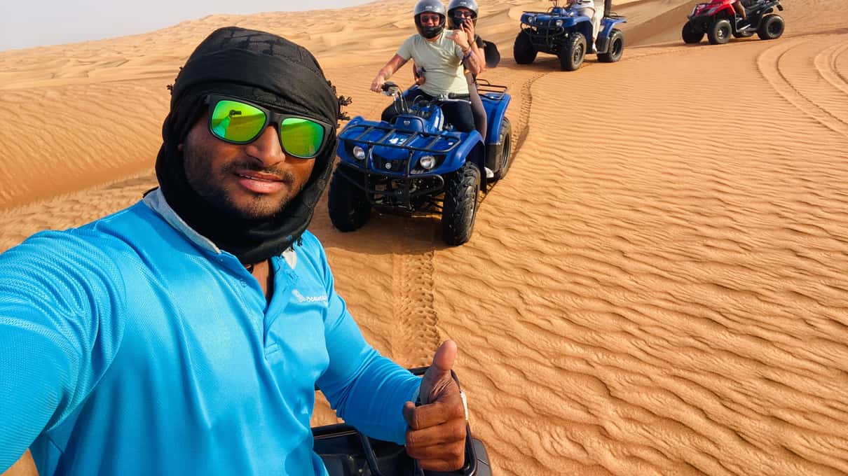 Guía con gafas de sol verdes y pañuelo se hace un selfi en las dunas mientras varios quads avanzan detrás de él