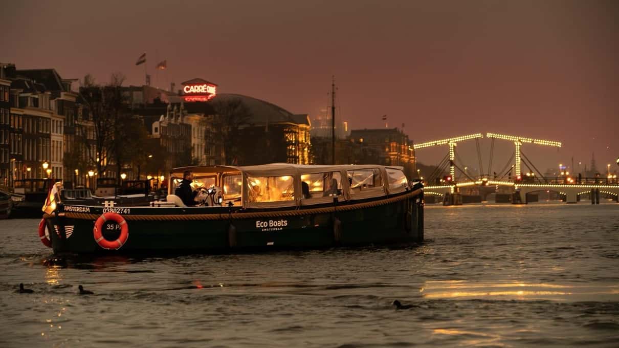 Barca elettrica di Eco Boats con la cabina illuminata sull’Amstel di sera, sullo sfondo la Magere Brug accesa