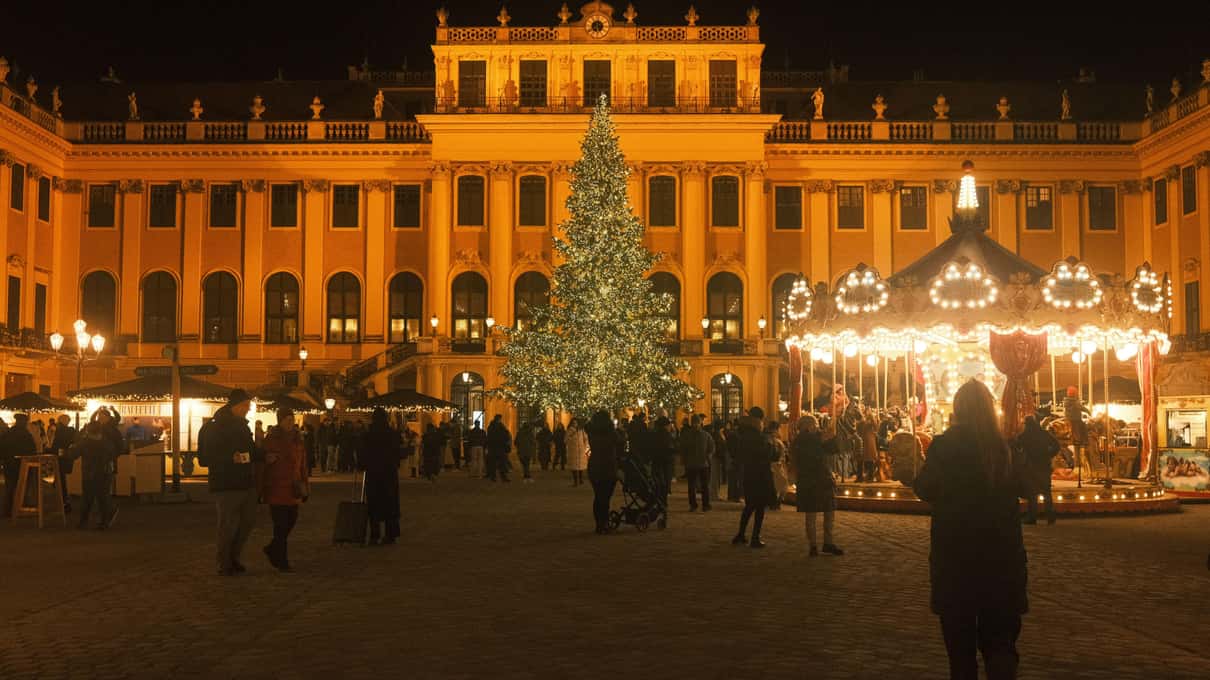 Schönbrunn Palace lit gold at night, with a tall Christmas tree, a lit carousel and a crowd of visitors in the forecourt market