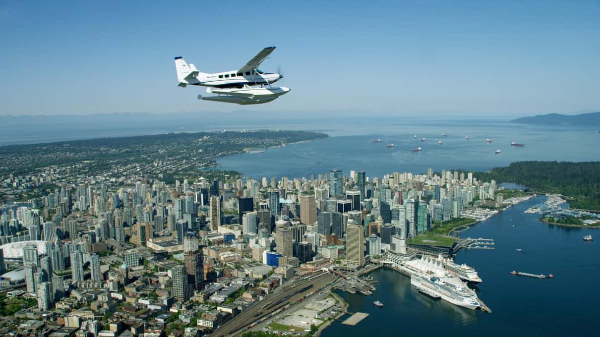 A Beaver floatplane flying over downtown Vancouver with cruise ships docked at Canada Place below
