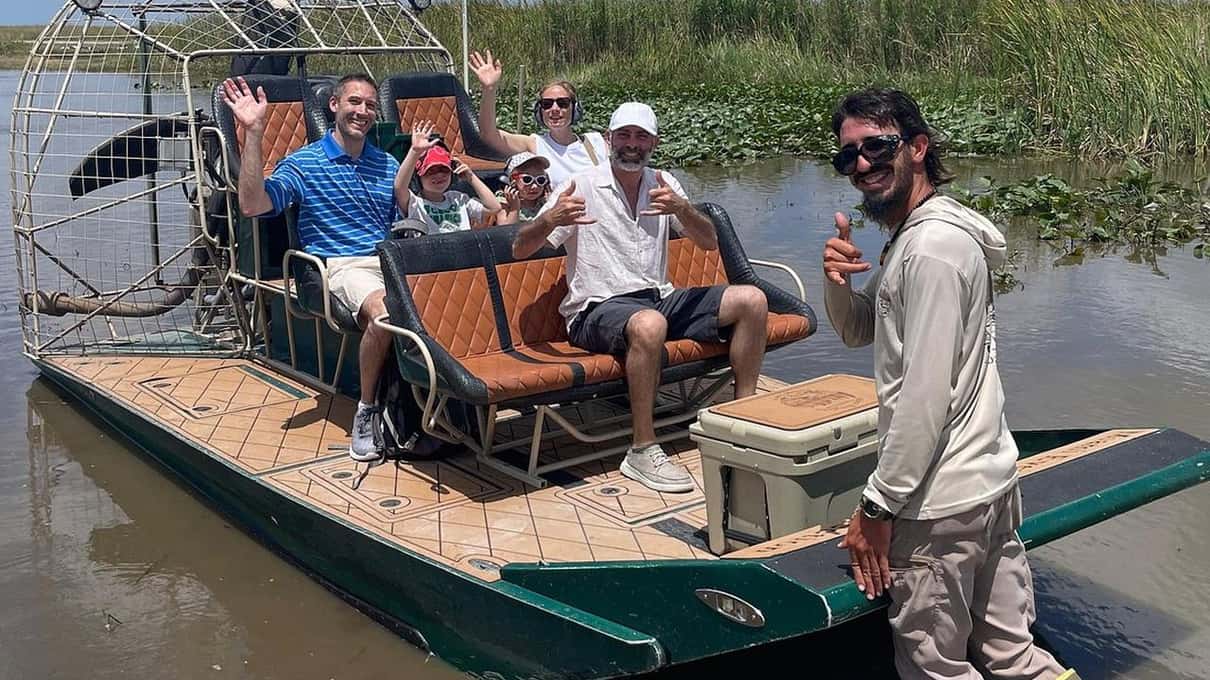 Guests waving from a small Miami Everglades Connection airboat while the captain stands in the shallow water beside it
