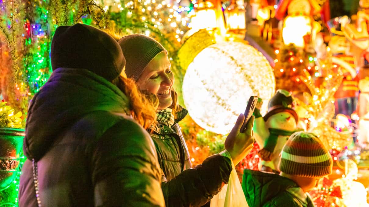 A visitor in a warm winter coat standing beside a giant glowing Christmas ornament in Dyker Heights